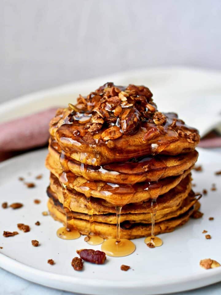 A stack of Pumpkin Spice Pancakes topped with pumpkin seed granola and maple syrup running down them.