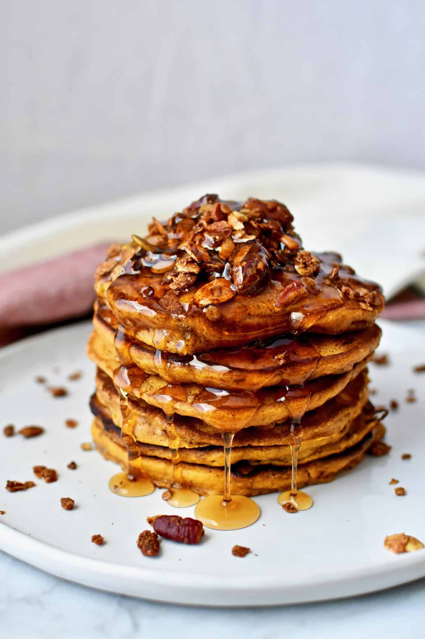 A stack of Pumpkin Spice Pancakes topped with pumpkin seed granola and maple syrup running down them.
