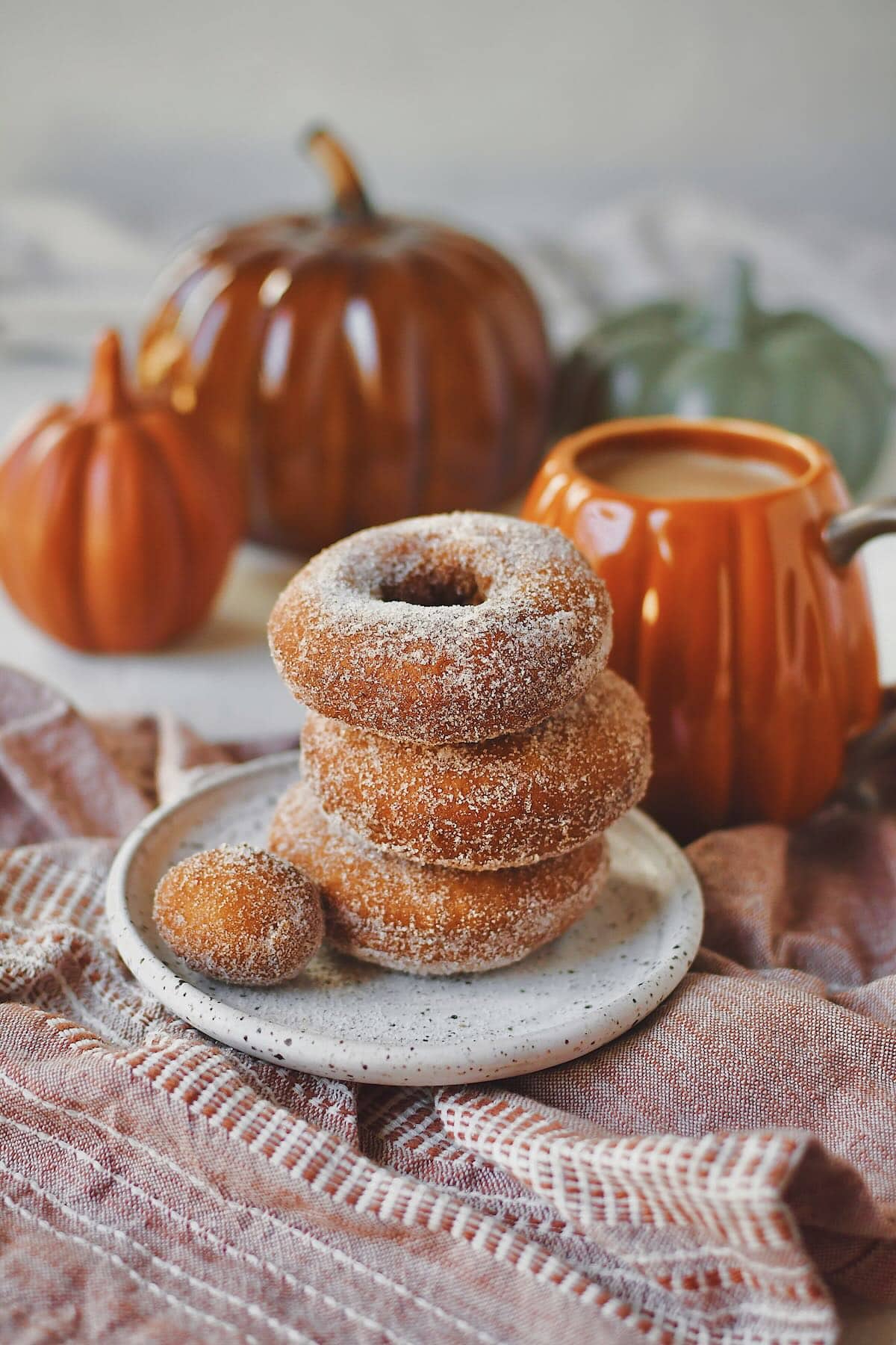 Pumpkin Donuts stacked up with a donut hole on the side, in front of a cup of coffee in a pumpkin mug.