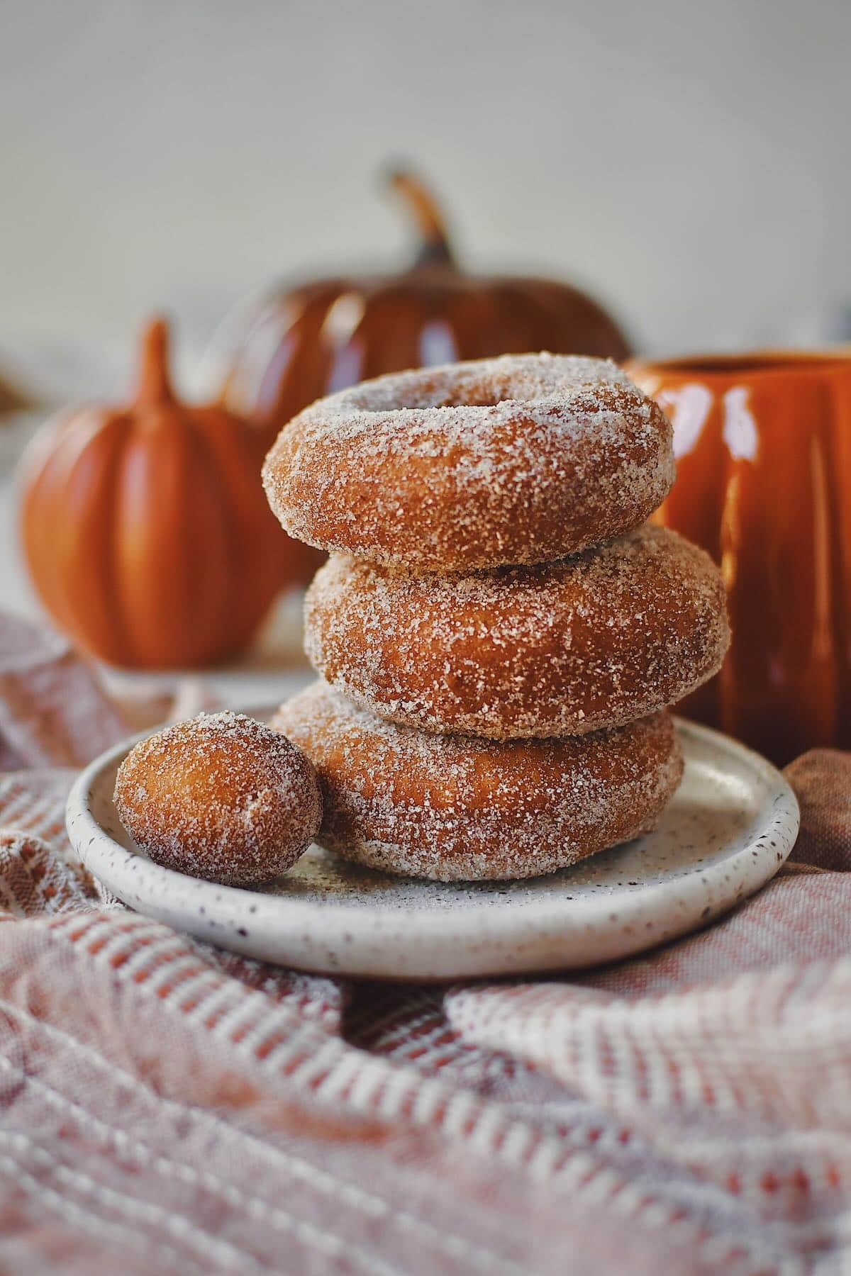 Pumpkin Donuts stacked up with a donut hole on the side, in front of a cup of coffee in a pumpkin mug.