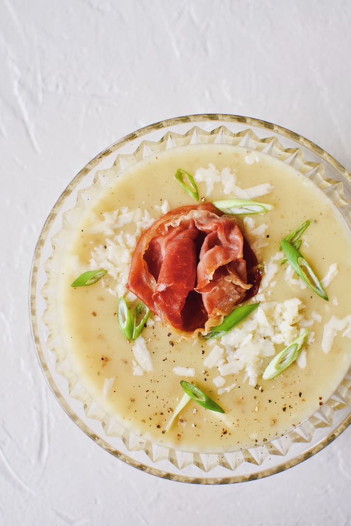 Creamy Potato Soup after blending, in a crystal bowl, topped with green onions, white cheddar, and a prosciutto rose.