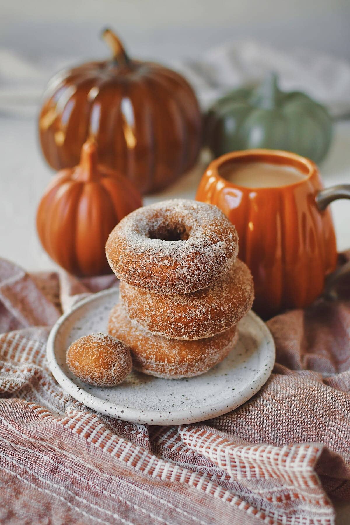 Pumpkin Donuts stacked up with a donut hole on the side, in front of a cup of coffee in a pumpkin mug.