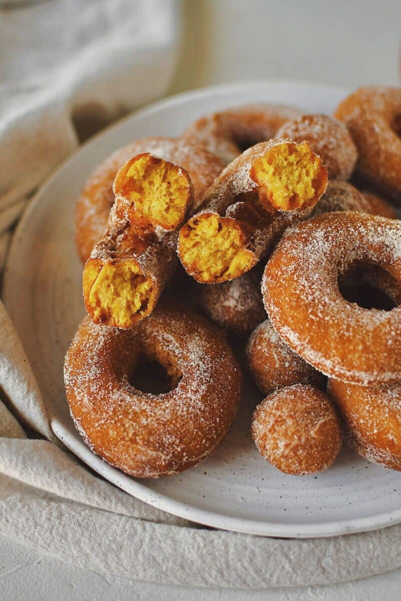 Pumpkin Donuts after frying, stacked on a plate and coating in pumpkin spice sugar, one broken in half to show the fluffy orange interior.