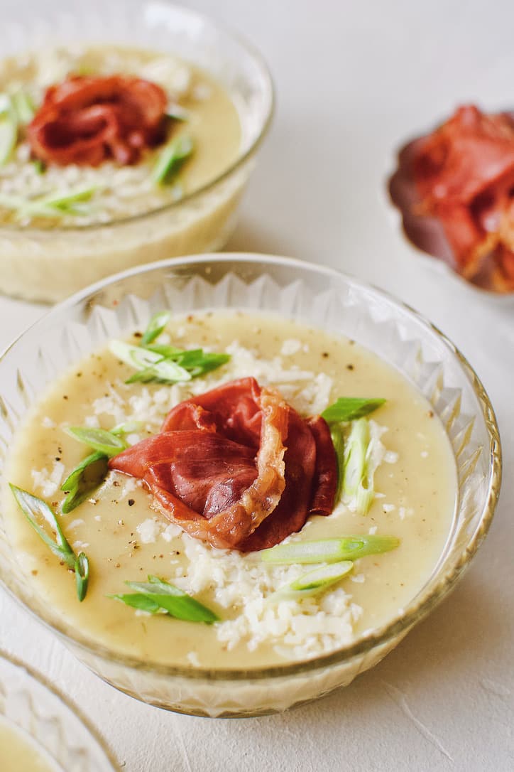 Creamy Potato Soup after blending, in a crystal bowl, topped with green onions, white cheddar, and a prosciutto rose.