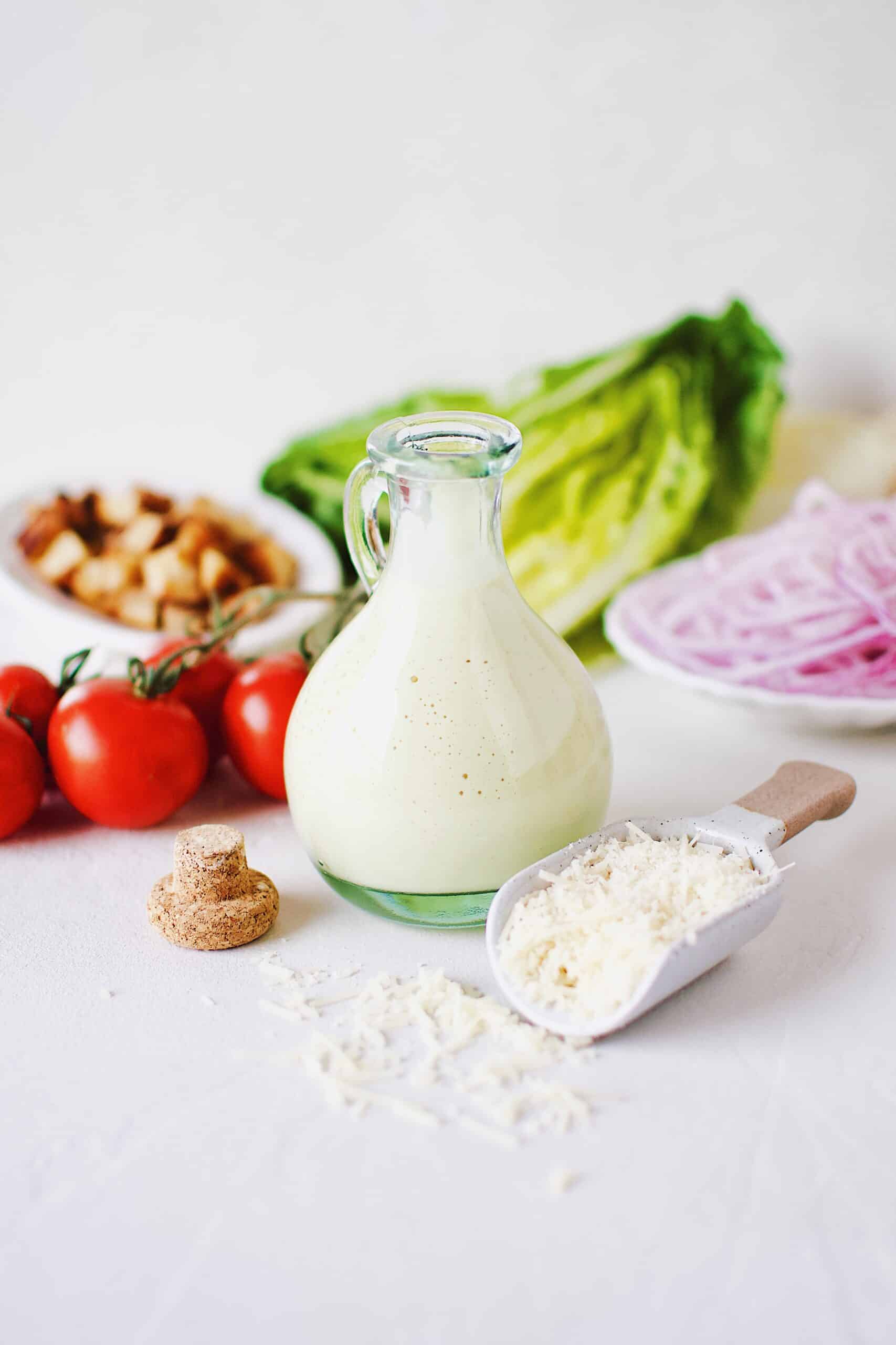 Creamy Caesar Dressing in a bottle surrounded by ingredients needed to make a Caesar Salad.