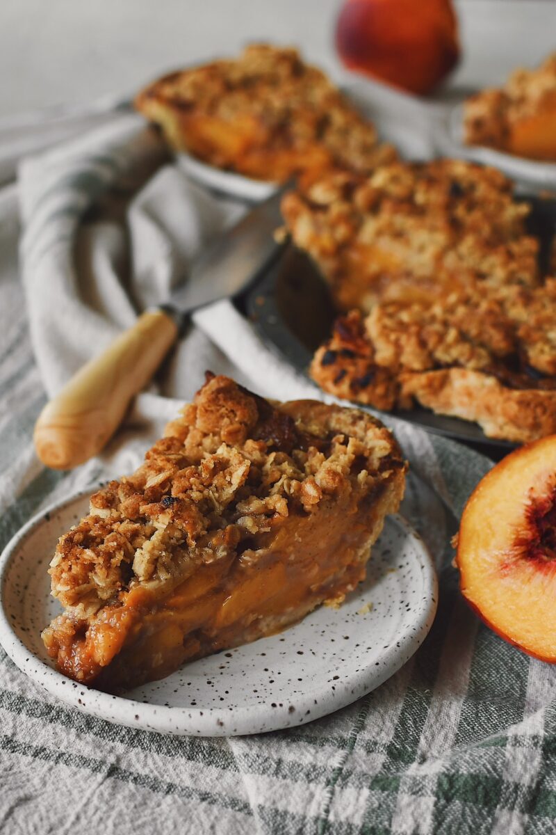 Looking at a slice of spiced peach pie, with a half peach on the side and remaining pie in the background.