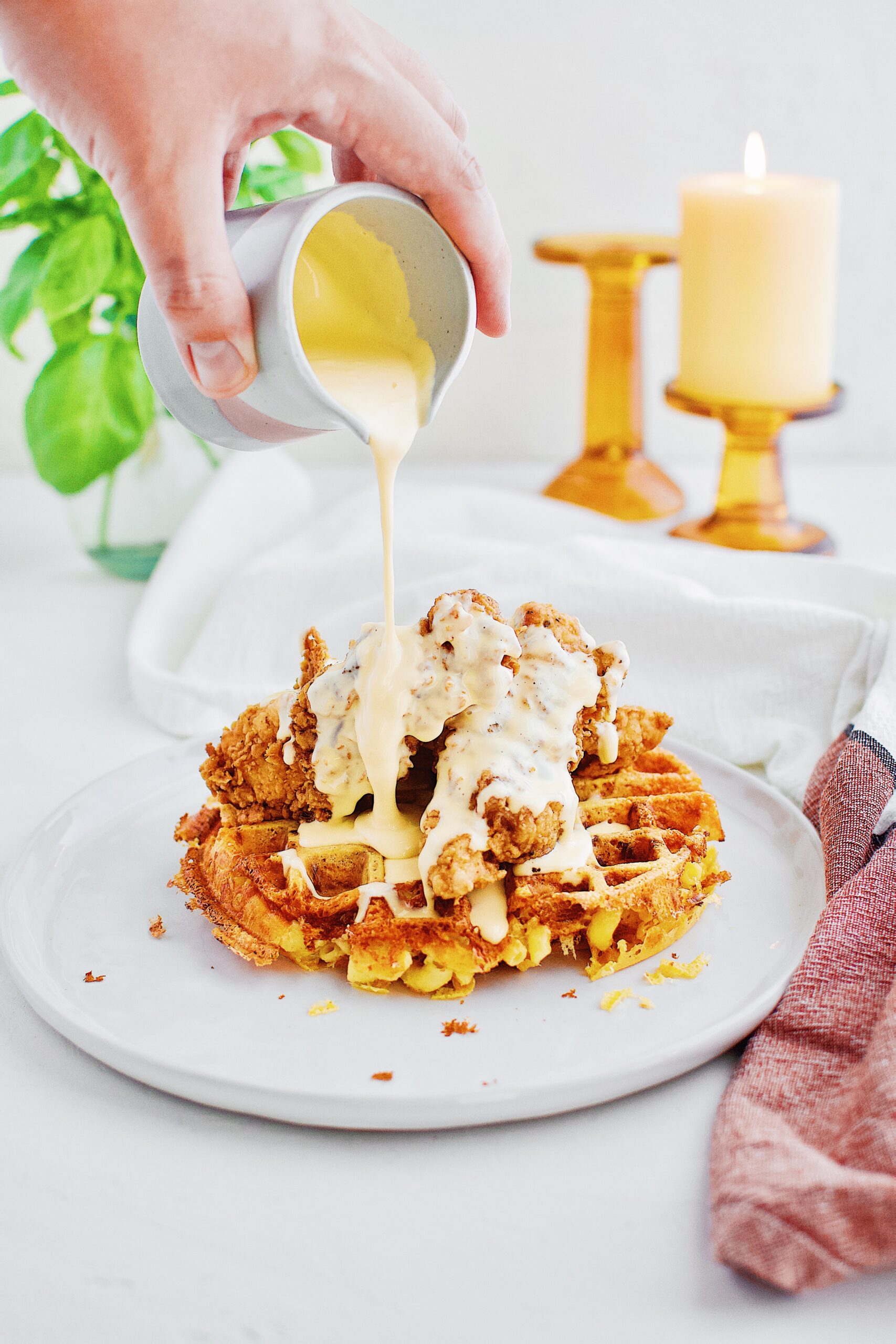 A hand pouring golden cheddar syrup over a stack of chicken mac n' cheese waffles on a white plate.