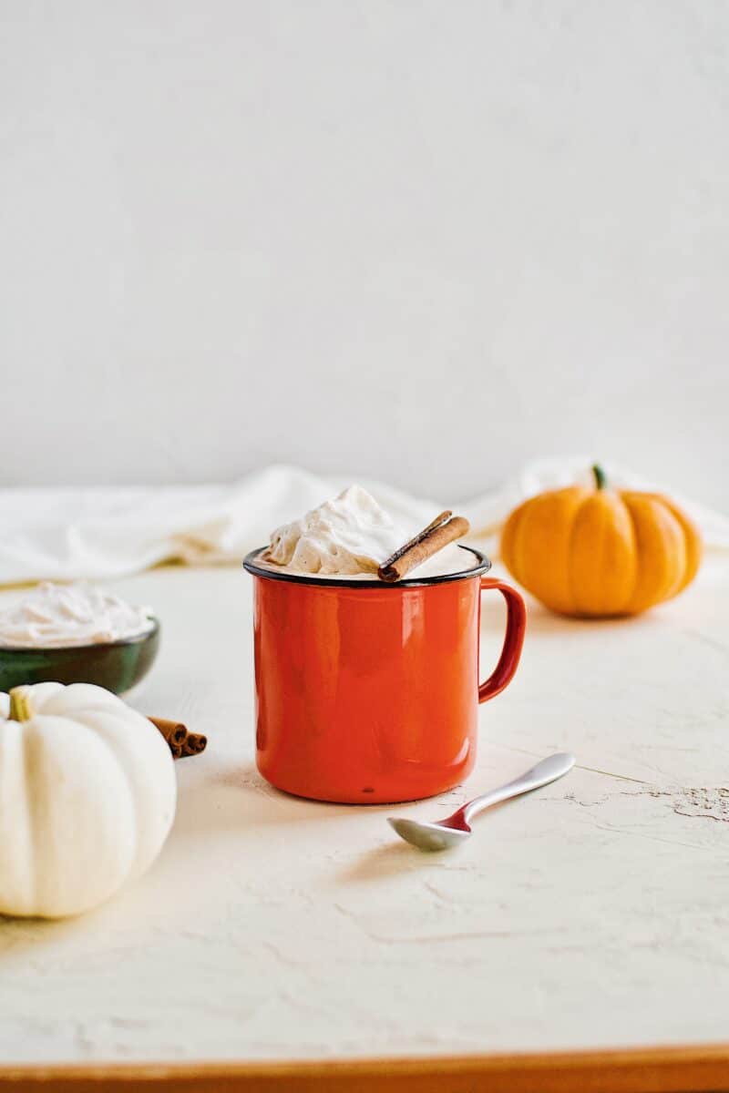 Pumpkin Spice Hot Chocolate in an orange camp mug topped with chai whipped cream and a cinnamon stick