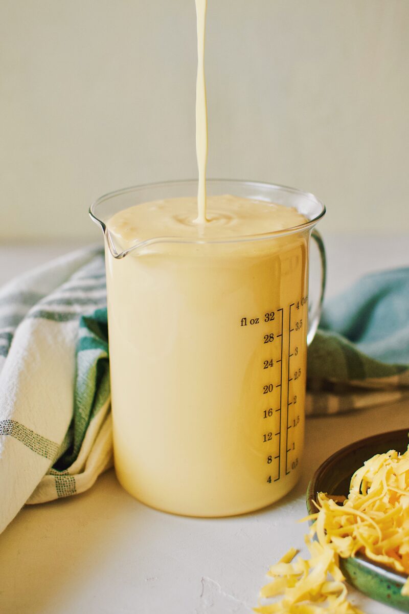 Golden cheddar syrup being poured from overhead into a glass measuring cup.