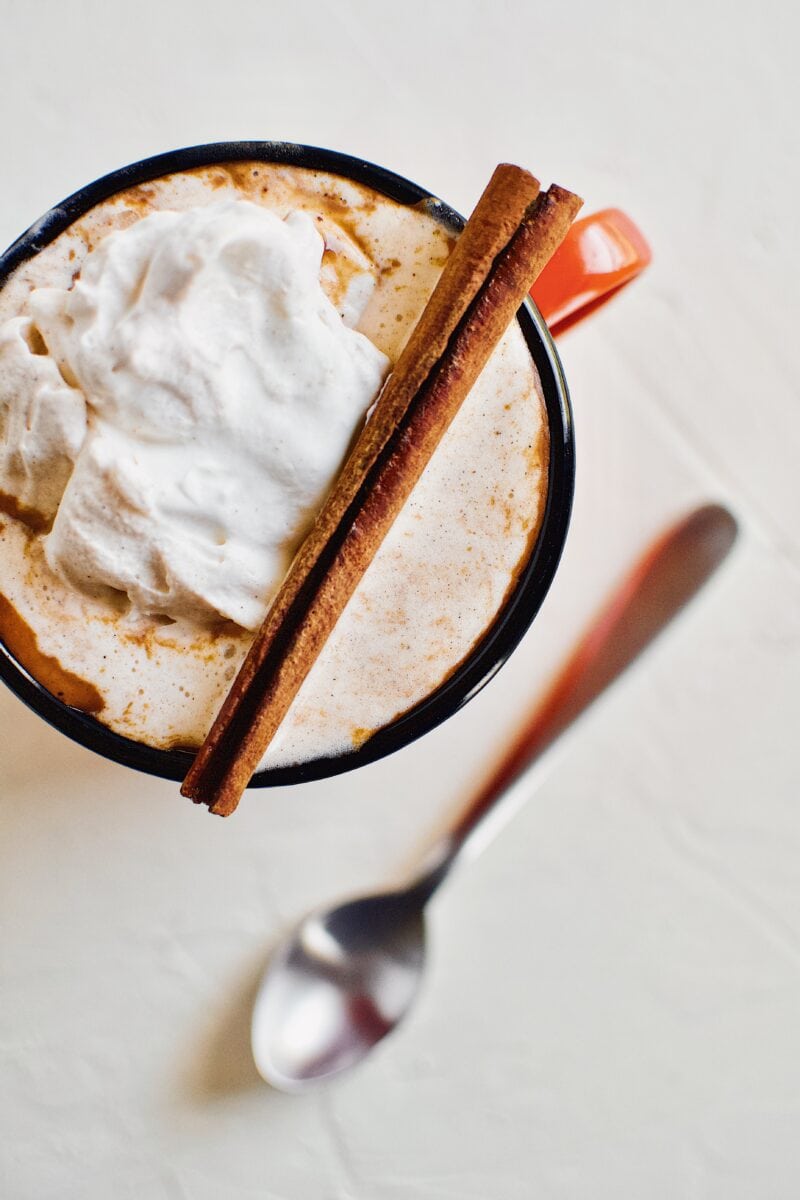 Pumpkin Spice Hot Chocolate in an orange camp mug topped with chai whipped cream and a cinnamon stick