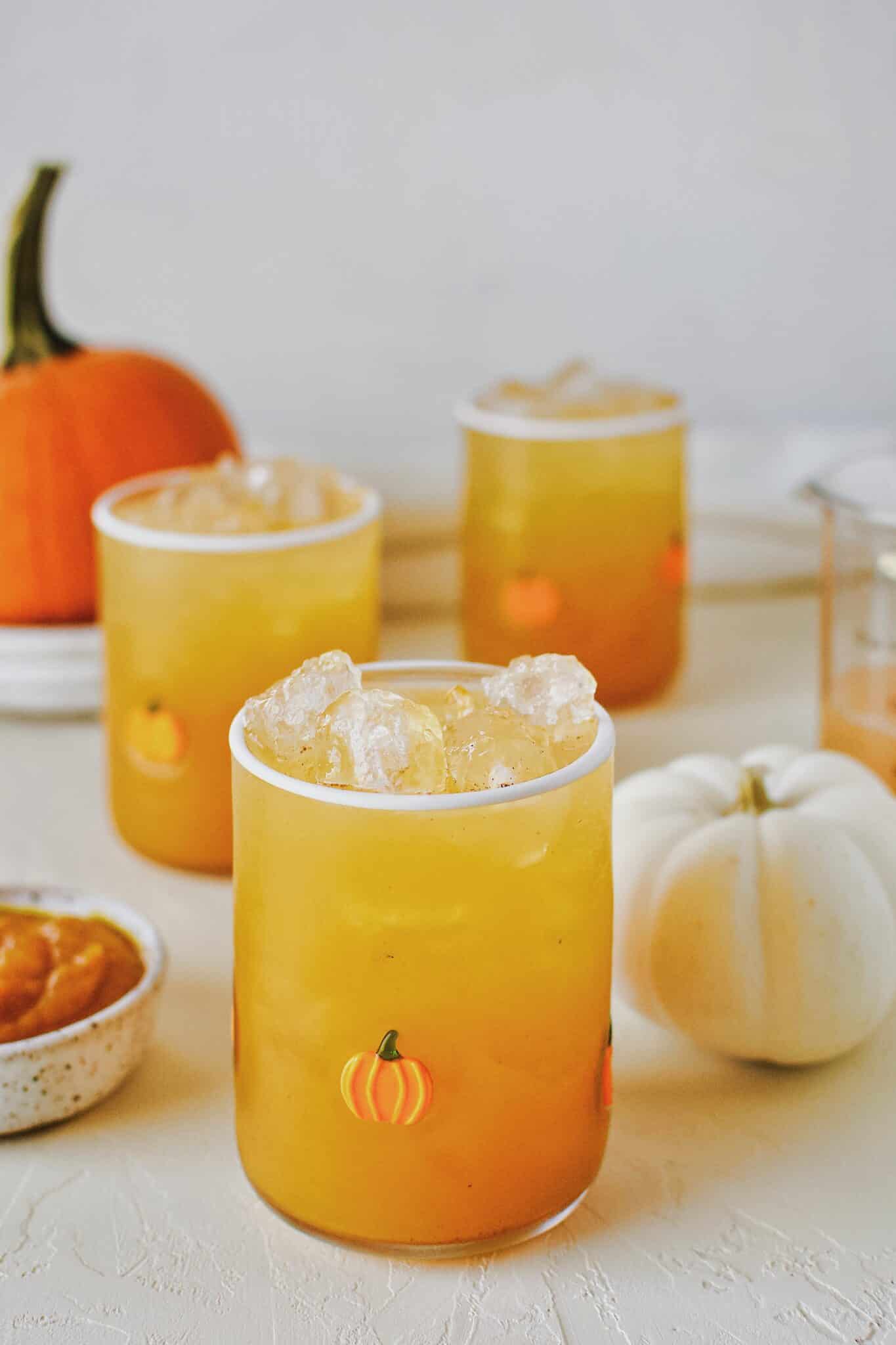 3 glasses of pumpkin juice in a row with baby pumpkin all around on a white background