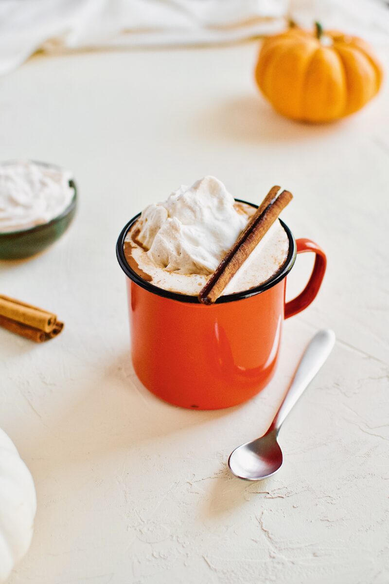 Pumpkin Spice Hot Chocolate in an orange camp mug topped with chai whipped cream and a cinnamon stick