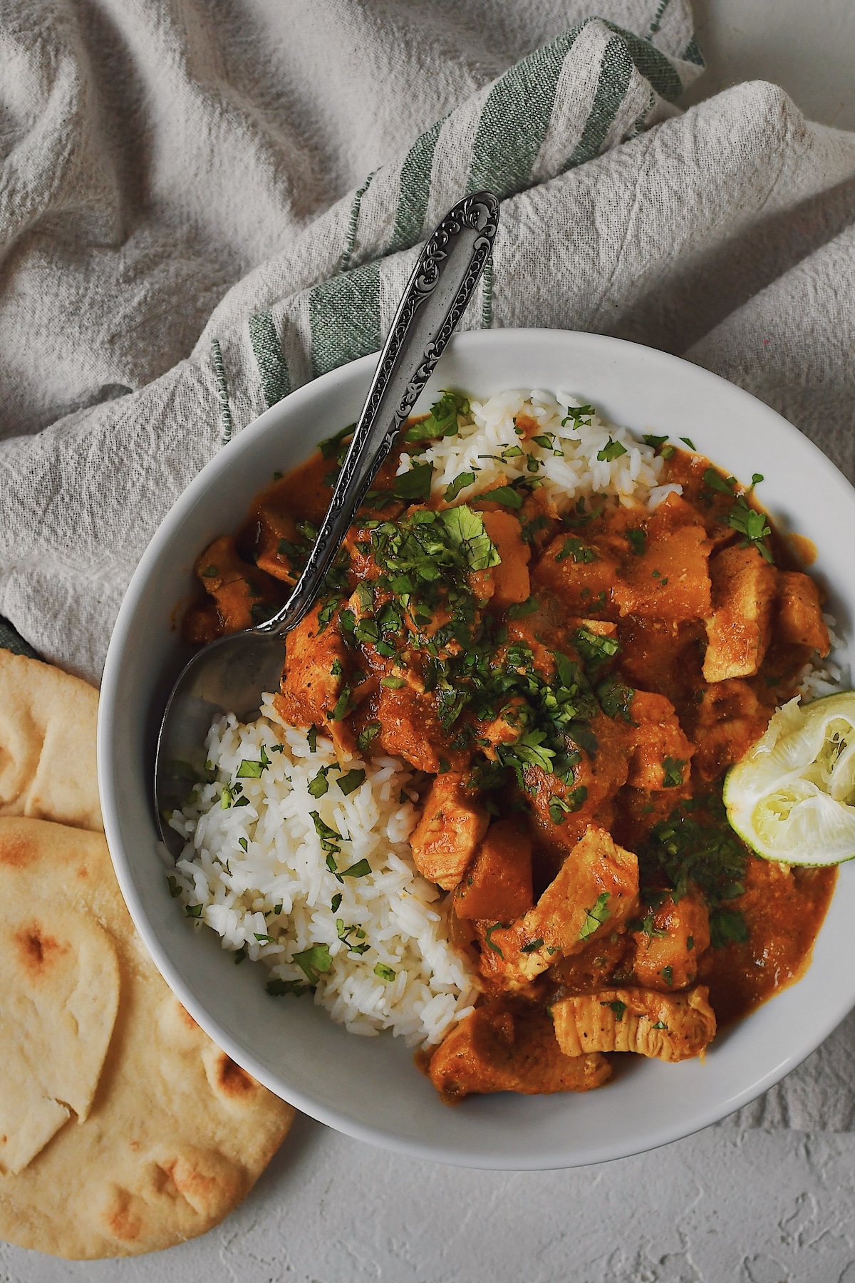 Looking down at a bowl of Pumpkin Butter Chicken served over rice with a lime wedge and cilantro. Some Naan Bread on the side.