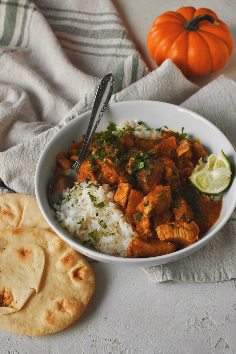 Looking into a bowl of Pumpkin Butter Chicken with a pumpkin in the background and naan bread served on the side.