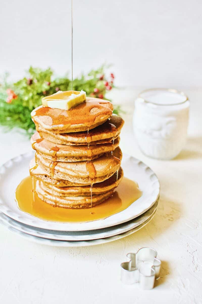 A stack of Gingerbread Pancakes topped with a pat of butter and maple syrup being drizzled over the top. A glass of milk sits in the corner in a santa shaped glass.