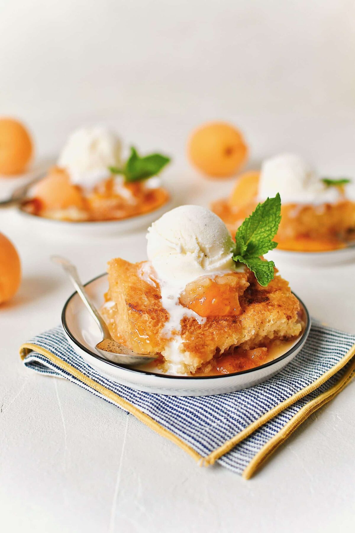 Apricot Cobbler served on small plates with a scoop of vanilla ice cream on top, a slice of fresh apricot, a sprig of mint, and a spoon to eat it with.