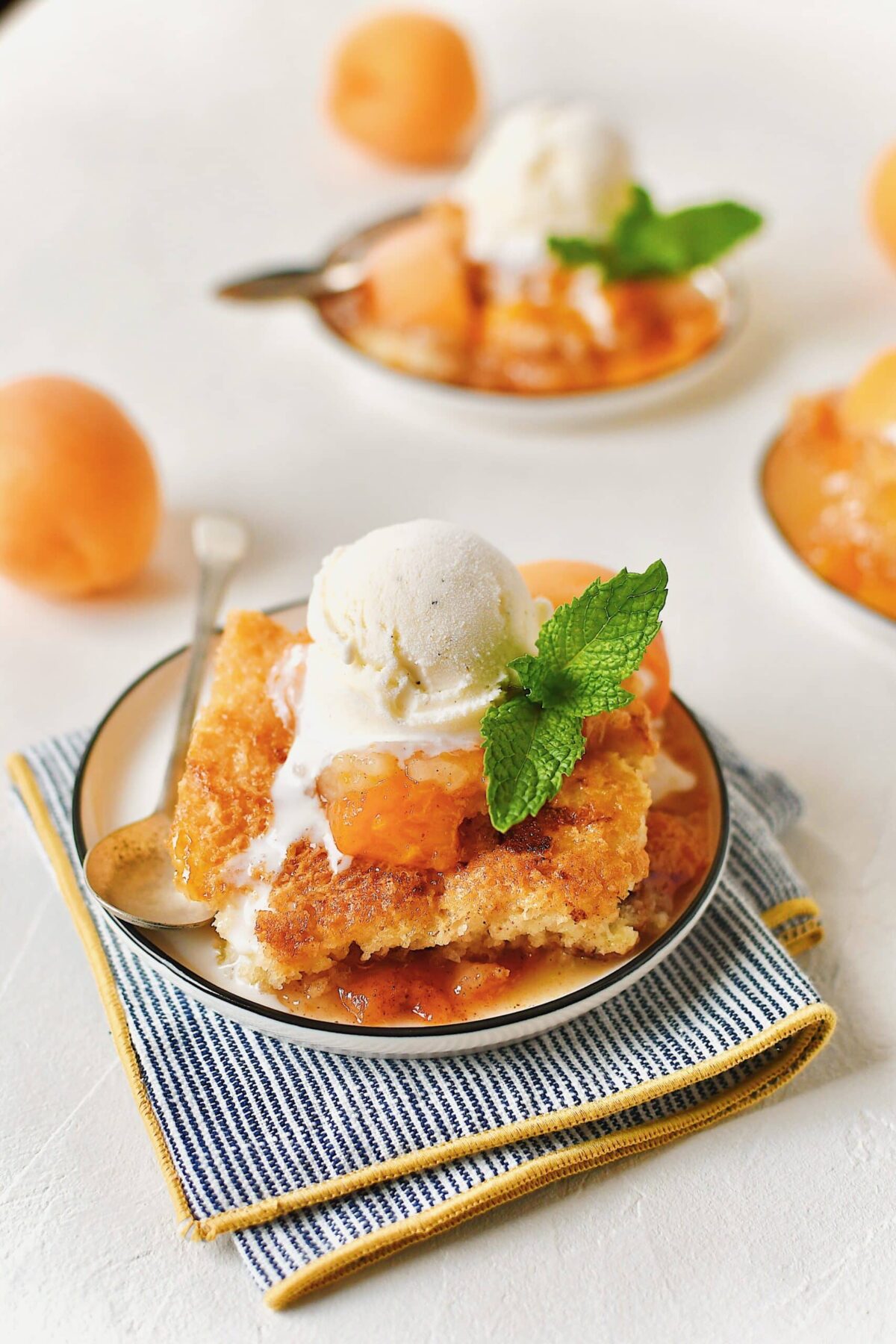 Apricot Cobbler served on small plates with a scoop of vanilla ice cream on top, a slice of fresh apricot, a sprig of mint, and a spoon to eat it with.