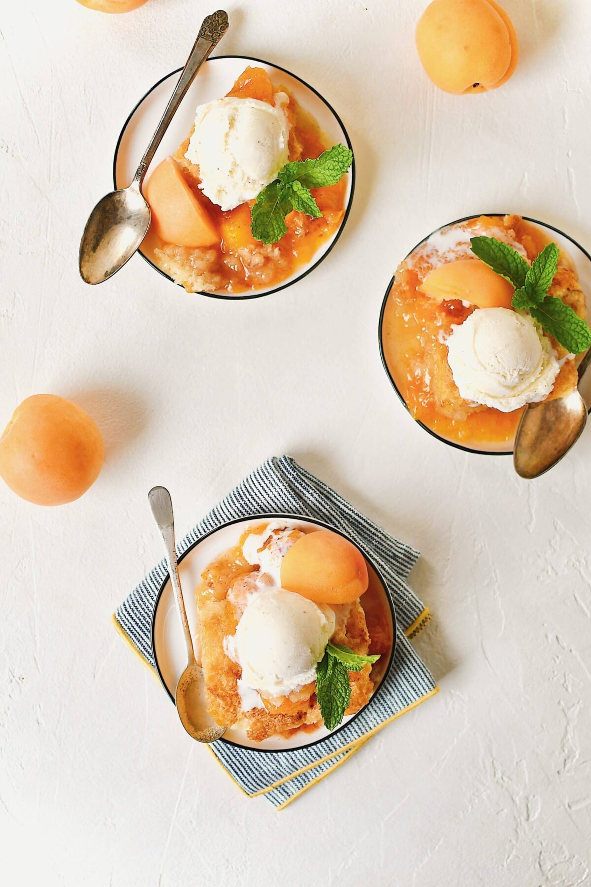 Apricot Cobbler served on small plates with a scoop of vanilla ice cream on top, a slice of fresh apricot, a sprig of mint, and a spoon to eat it with.