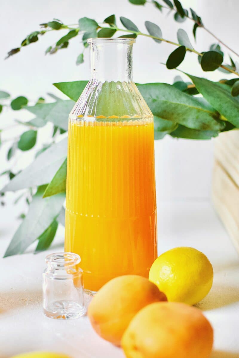 Apricot Nectar Juice in a glass with ice, surrounded by fresh apricots and lemons, with a pitcher of apricot juice in the background.