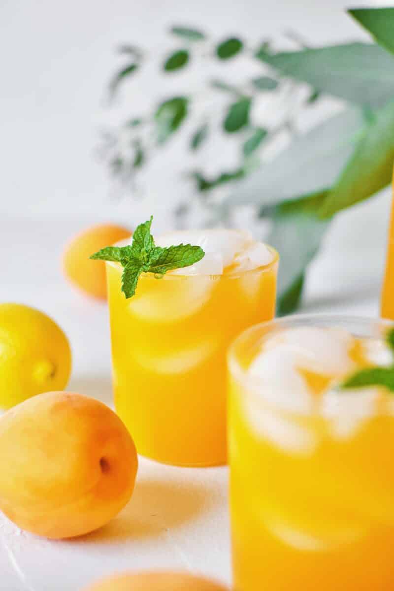 Apricot Nectar Juice in a glass with ice, surrounded by fresh apricots and lemons, with a pitcher of apricot juice in the background.