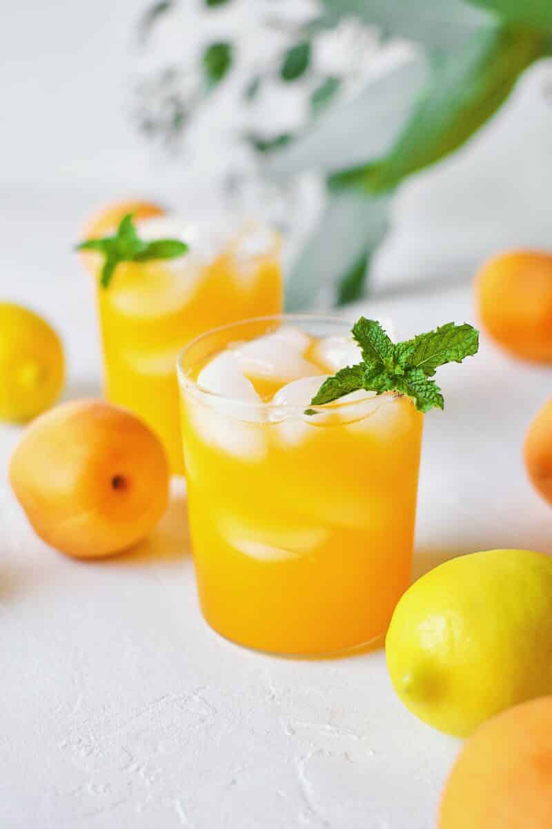 Apricot Nectar Juice in a glass with ice, surrounded by fresh apricots and lemons, with a pitcher of apricot juice in the background.