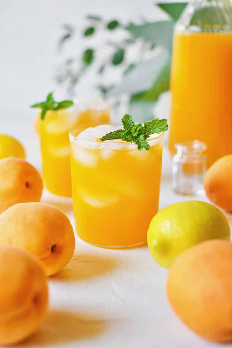 Apricot Nectar Juice in a glass with ice, surrounded by fresh apricots and lemons, with a pitcher of apricot juice in the background.