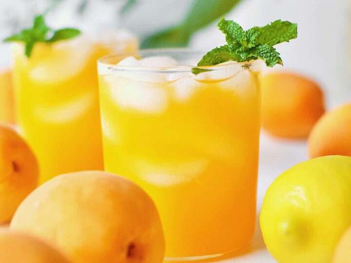 Apricot Nectar Juice in a glass with ice, surrounded by fresh apricots and lemons, with a pitcher of apricot juice in the background.