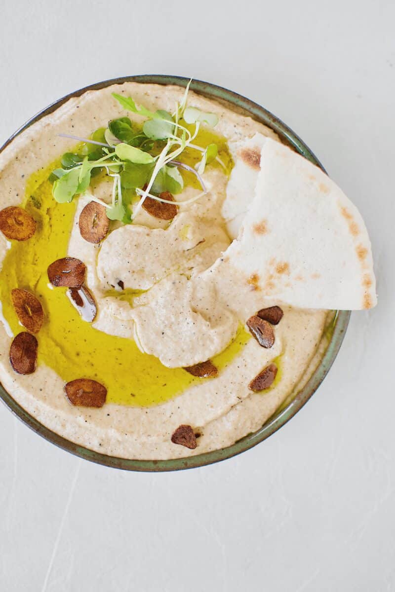 Baba Ghanoush with Toasted Garlic in a bowl, topped with olive oil and sliced toasted garlic. And a piece of pita dipped in.