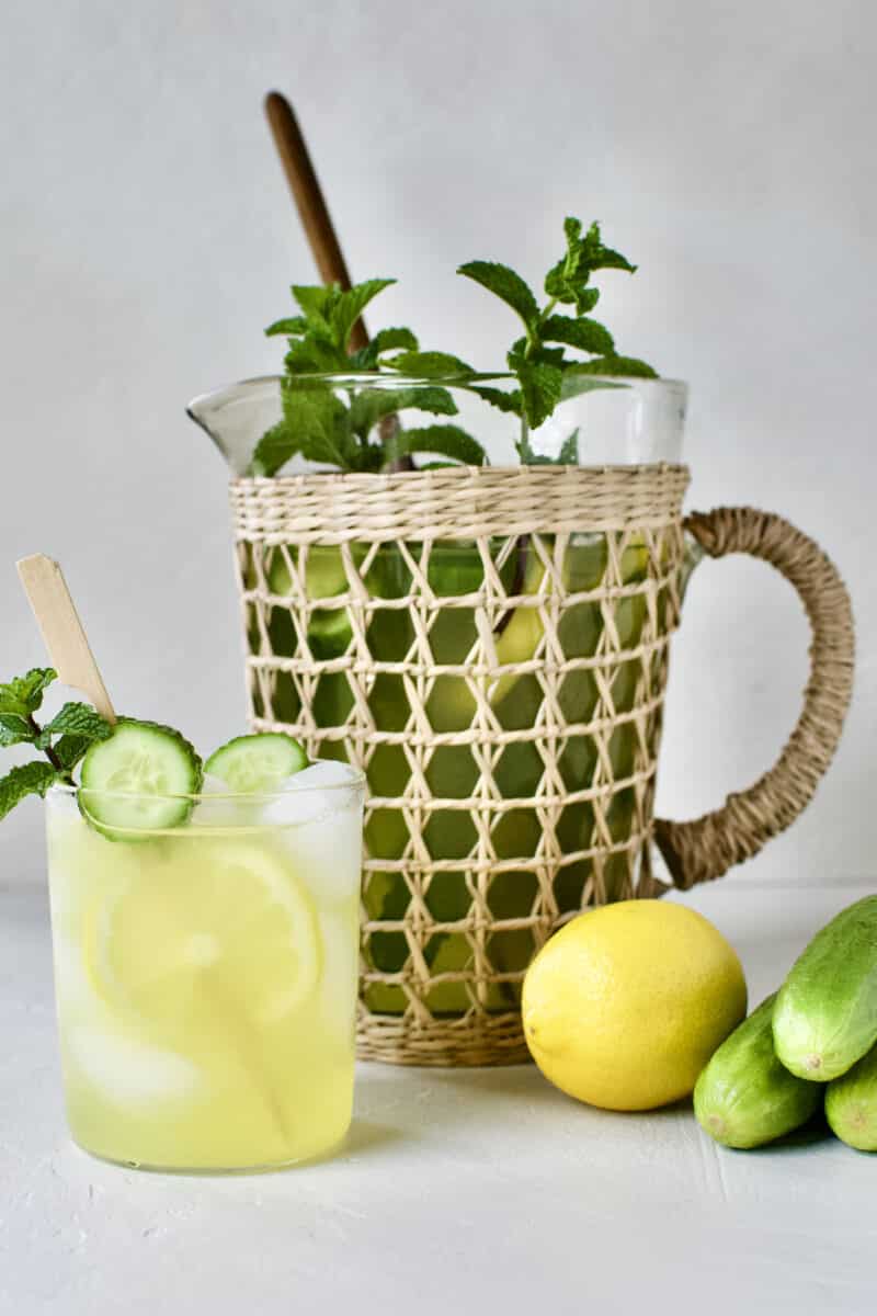 Cucumber Mint Lemonade in a glass with a skewer of lemon and cucumber slices and a sprig of mint. A pitcher of lemonade behind the glass.