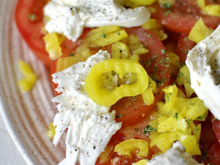 Tomato slices on a platter, topped with chopped banana pepper pieces, banana pepper vinaigrette, and burrata cheese.