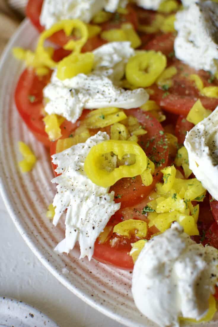 Tomato slices on a platter, topped with chopped banana pepper pieces, banana pepper vinaigrette, and burrata cheese.