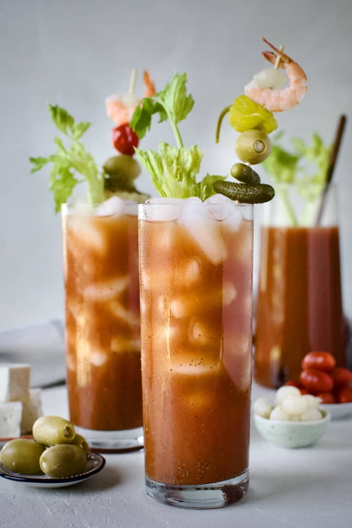 Bloody Mary's ready to drink, topped with things like pickles, olives, peppers, and shrimp. With a pitcher of mix in the background.