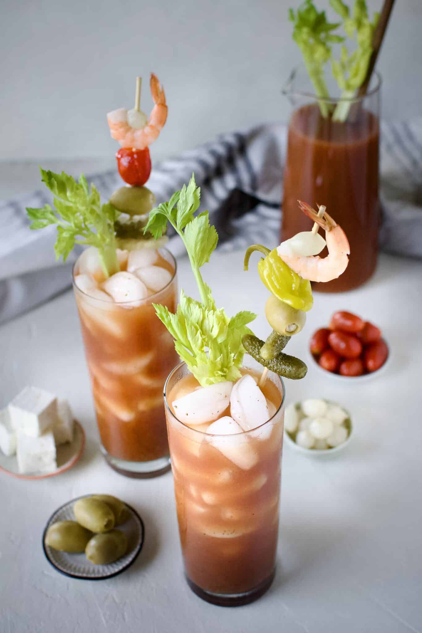 Bloody Mary's ready to drink, topped with things like pickles, olives, peppers, and shrimp. With a pitcher of mix in the background.