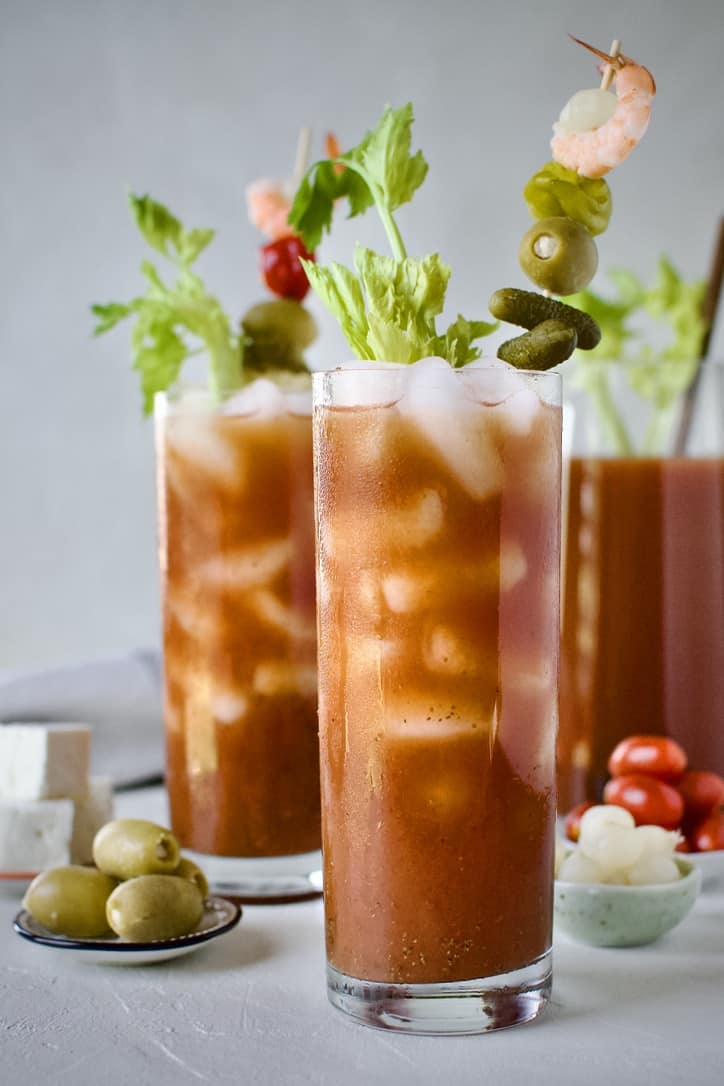 Bloody Mary's ready to drink, topped with things like pickles, olives, peppers, and shrimp. With a pitcher of mix in the background.