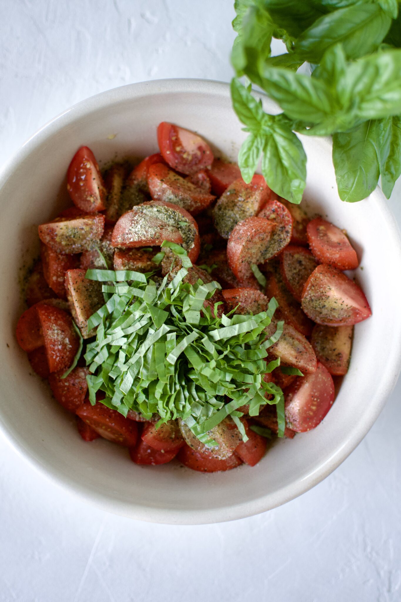 Seasoned tomatoes in a large bowl with olive oil, seasonings, vinegar, and basil strands.