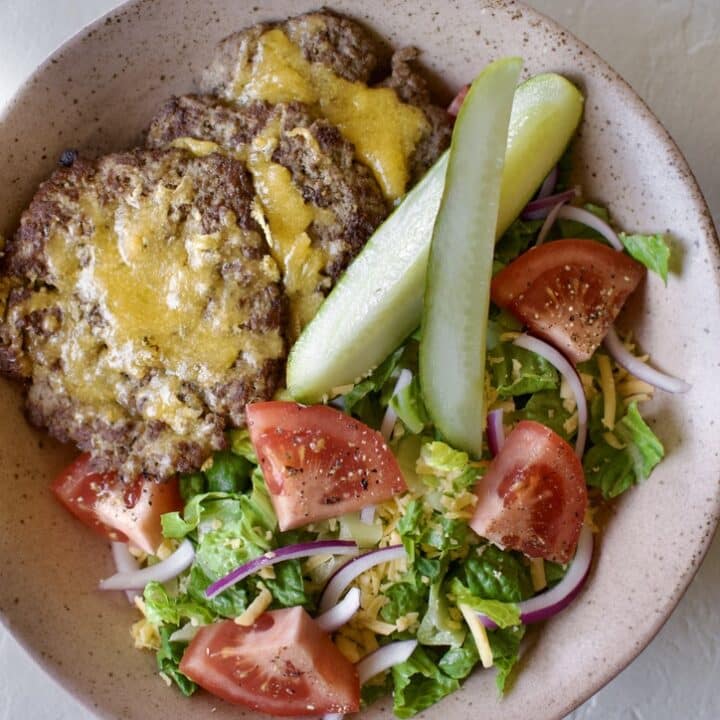 Lettuce in a bowl, topped with cheddar cheese , red onions, tomatoes, pickles, & cheeseburger patties.