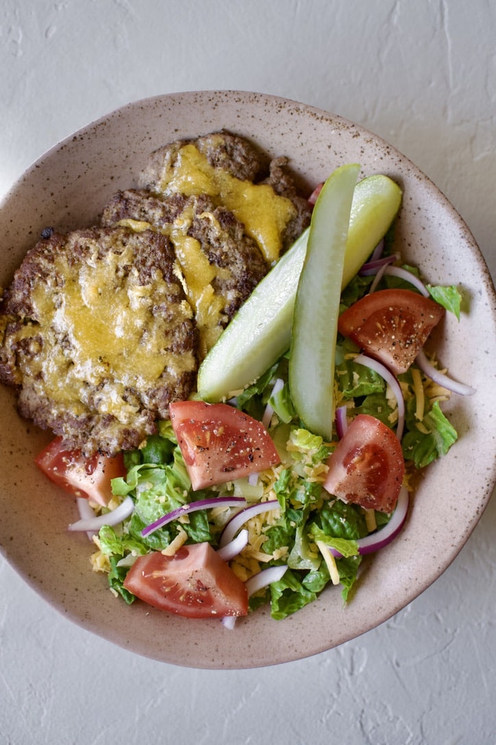Lettuce in a bowl, topped with cheddar cheese , red onions, tomatoes, pickles, & cheeseburger patties.