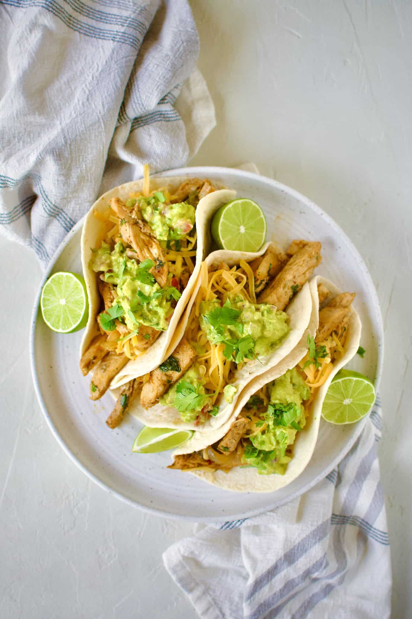 Easy Chicken Fajitas in tortillas topped with cheddar cheese, guacamole, and cilantro.