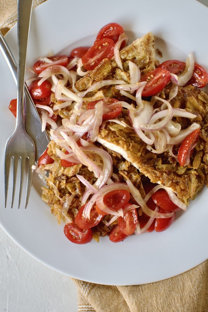 Sliced potato chip chicken on a plate topped with pickled onions and tomatoes.