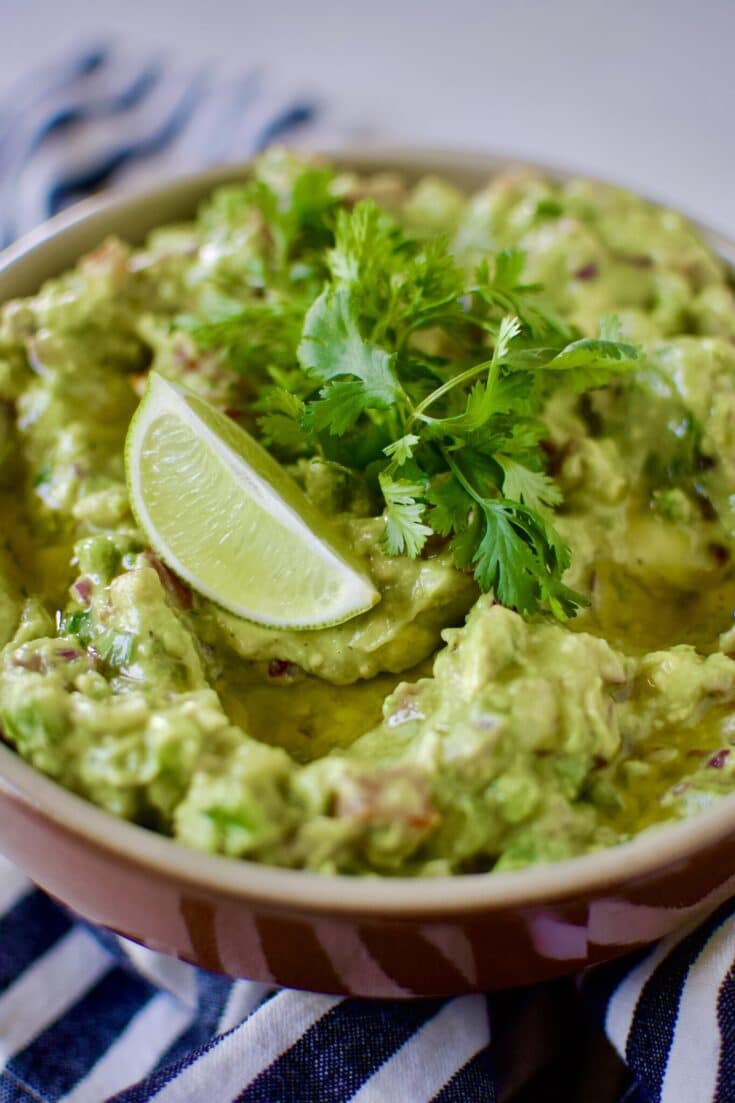 Mexican Guacamole Recipe in a bowl topped with a lime wedge, baby cilantro sprigs and olive oil.