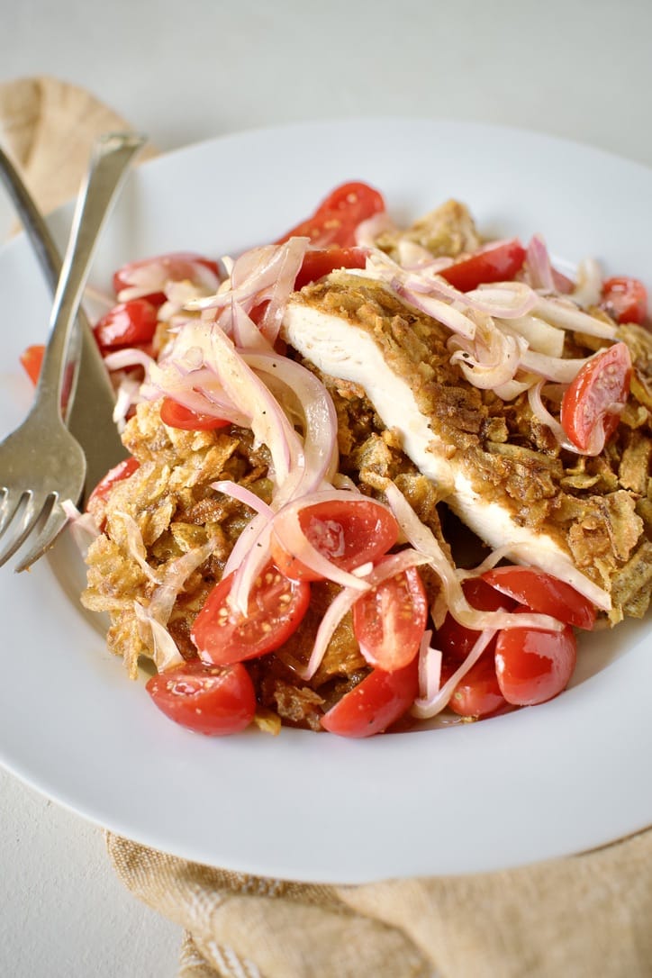 Sliced potato chip chicken on a plate topped with pickled onions and tomatoes.