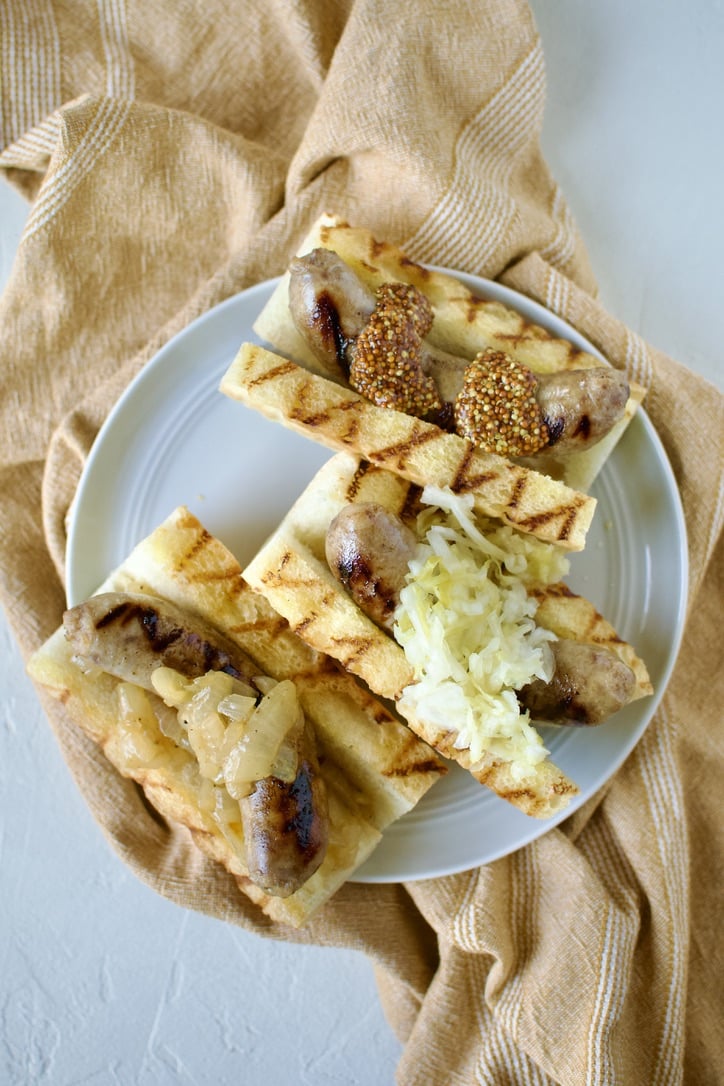 Braised and Grilled Brats served on grilled buns one with onions on it, one with sauerkraut, one with mustard.