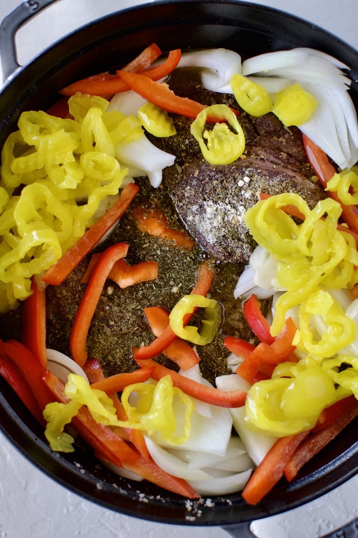 Seared beef chuck roast in a pot with onions, bell peppers, seasonings and pepperoncini ready to stew.