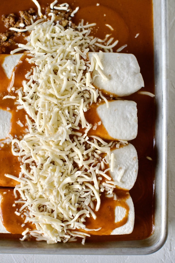Rolled enchiladas in a baking pan, topped with red enchilada sauce, and cheese. Before baking.