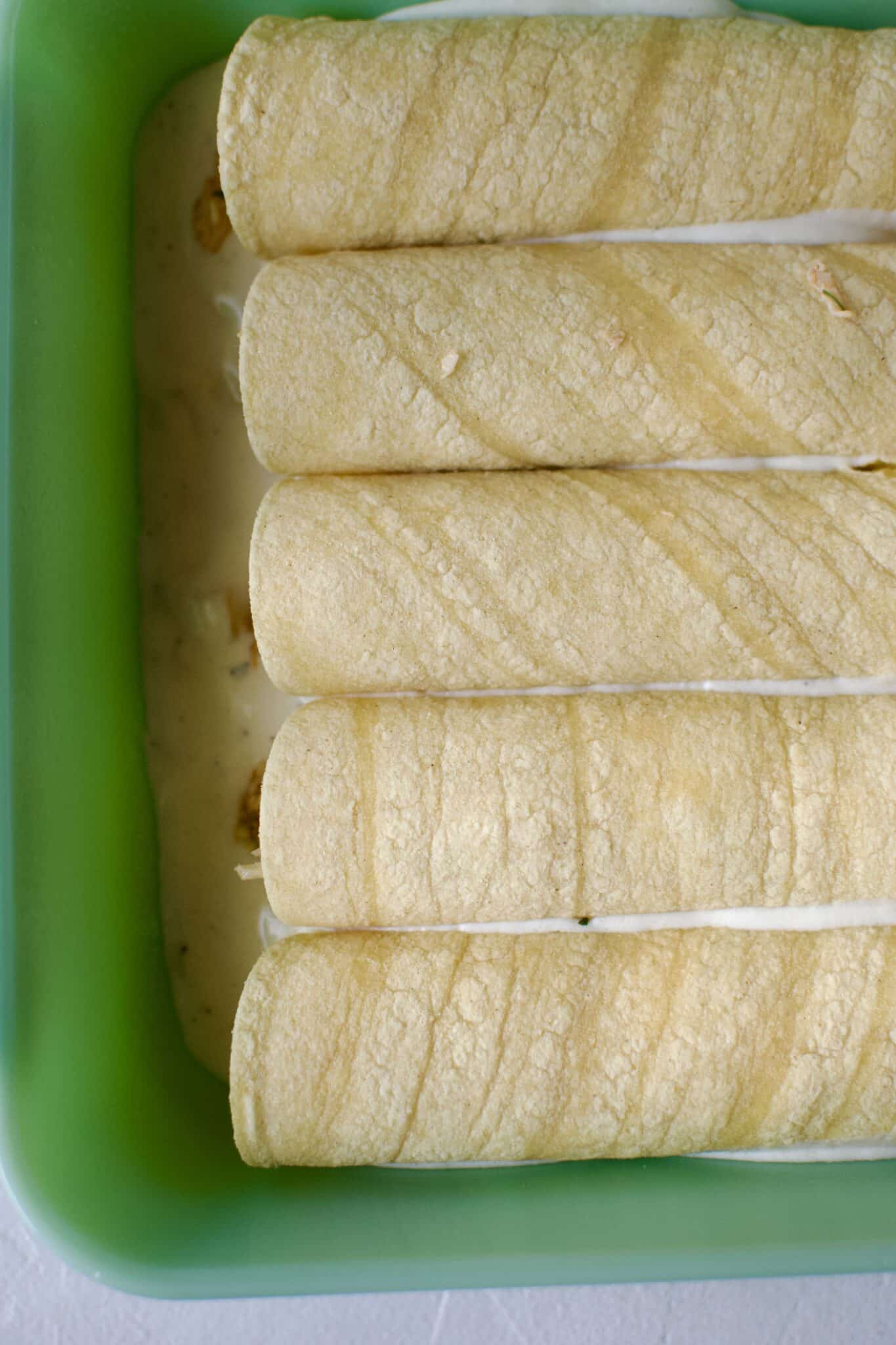 Chicken filled tortillas rolled up and placed in a baking dish that has been lined with sour cream enchilada sauce.