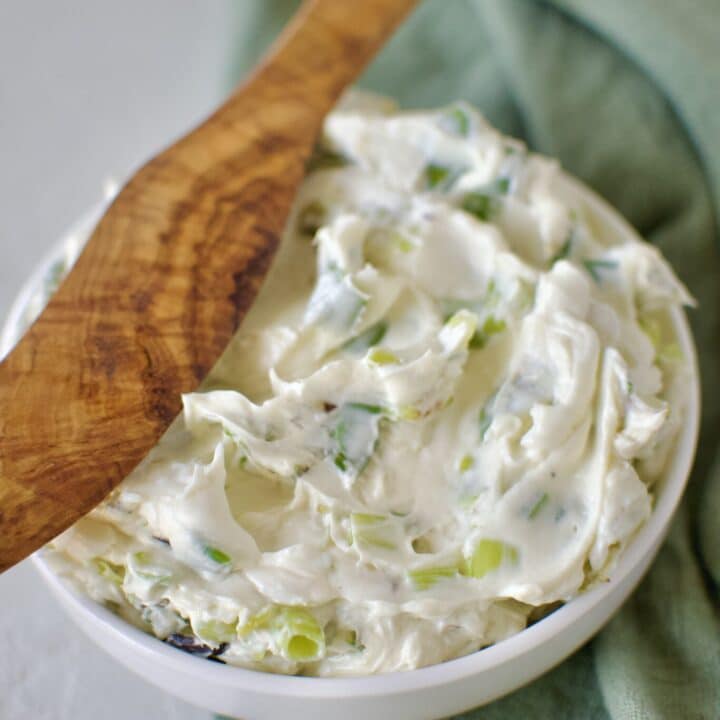 Finished scallion cream cheese in a dish ready to eat. With a wood spreading knife resting on the side.