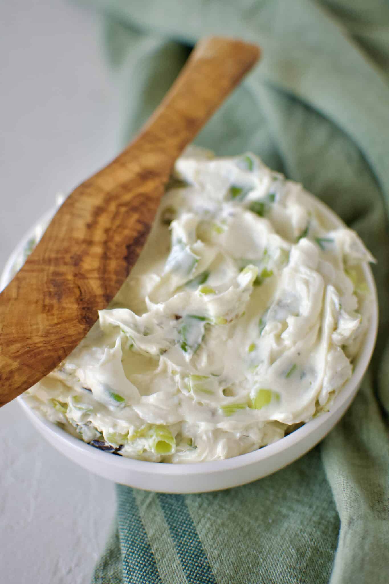 Finished scallion cream cheese in a dish ready to eat. With a wood spreading knife resting on the side.