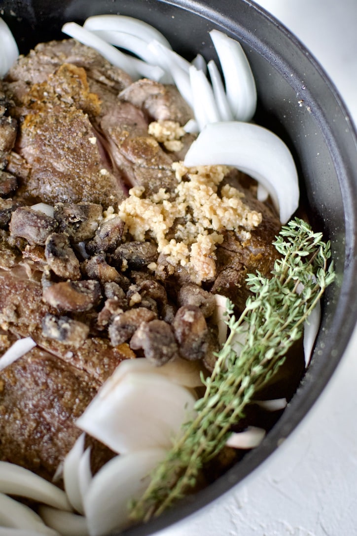 Seared chuck roast in a large pot with onions, cooked mushrooms, garlic, thyme, red wine, and beef stock.