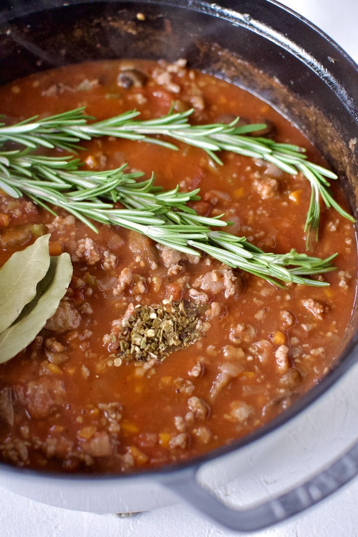 Adding tomatoes, bay leaves, rosemary sprigs, and crushed pepper flakes to the cooked veggies and sausage in the pot.