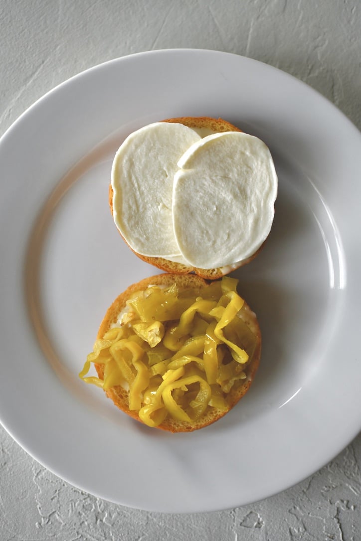 Assembling a roast beef sandwich; placing the banana peppers and mozzarella cheese onto the toasted bun that has horseradish sauce on it.